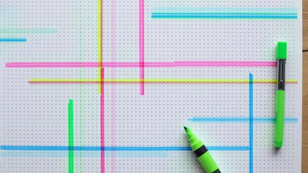 Overhead view of a sheet of paper covered in a dotted grid, crossed by pink, yellow, blue and green highlighter stripes that run horizontally and vertically, with a green highlighter pen resting on the grid.