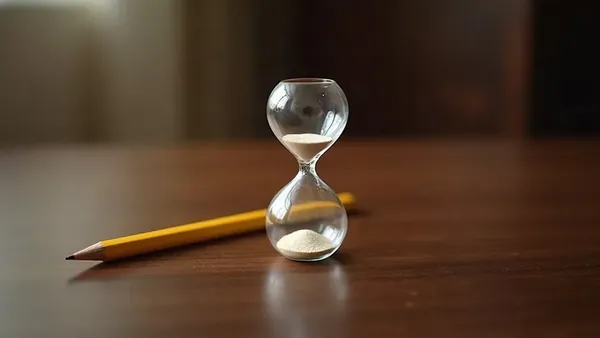 A sharpened yellow pencil lying beside a glass sand hourglass on a dark wooden desk in warm light